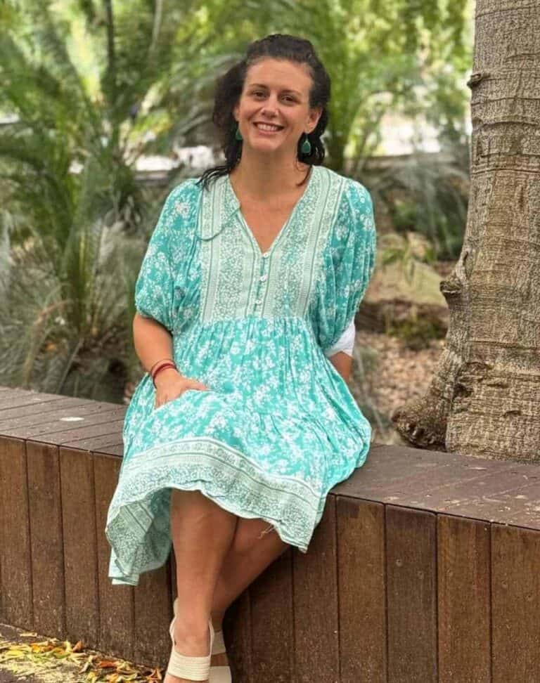 A lady smiles sitting on a park bench, looking into the camera.